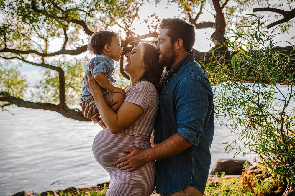 Family maternity photo by the water during golden hour with autumn trees in background.