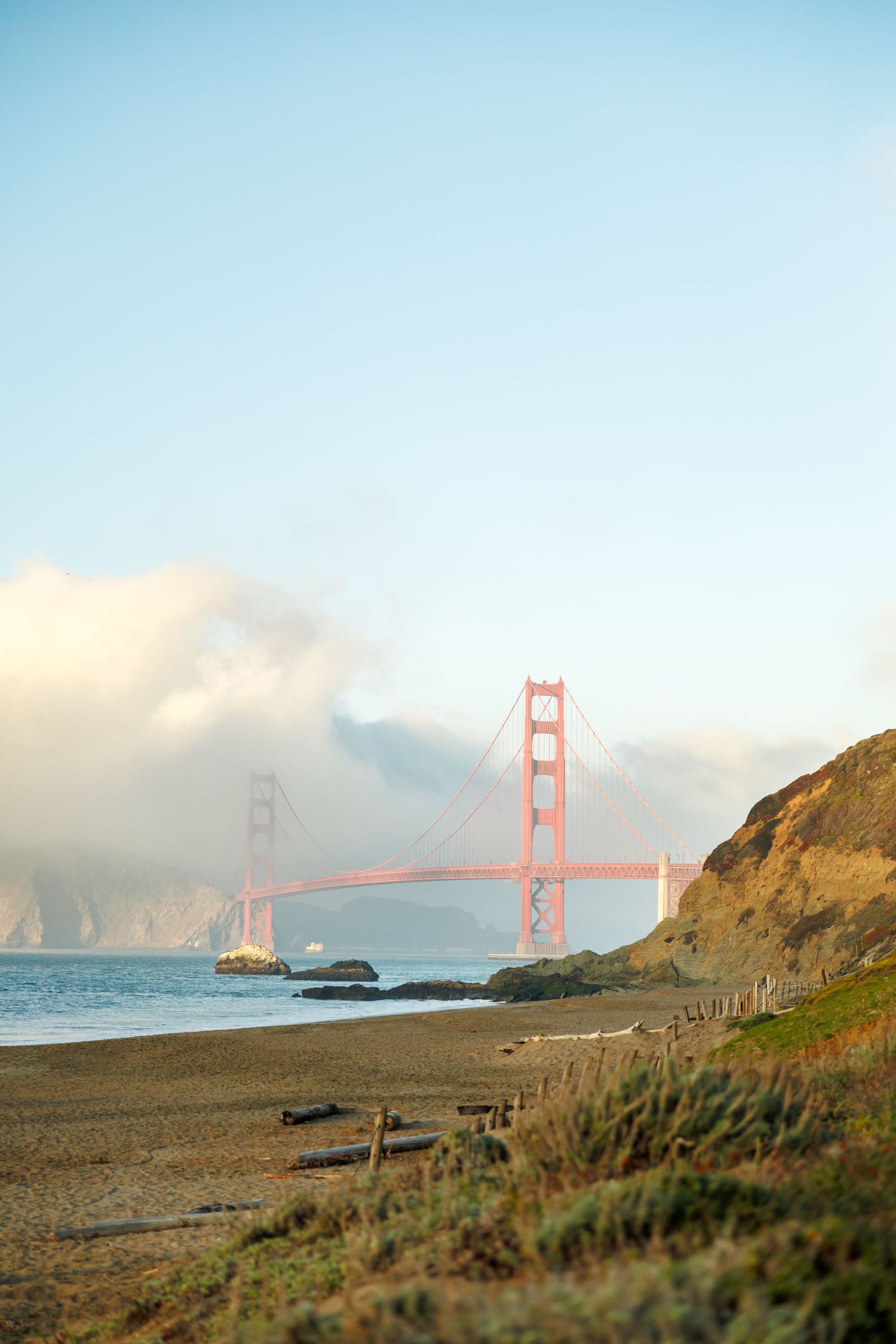 The Golden Gate Bridge emerges from foggy clouds along San Francisco's coastline with beach and cliffs in the foreground.