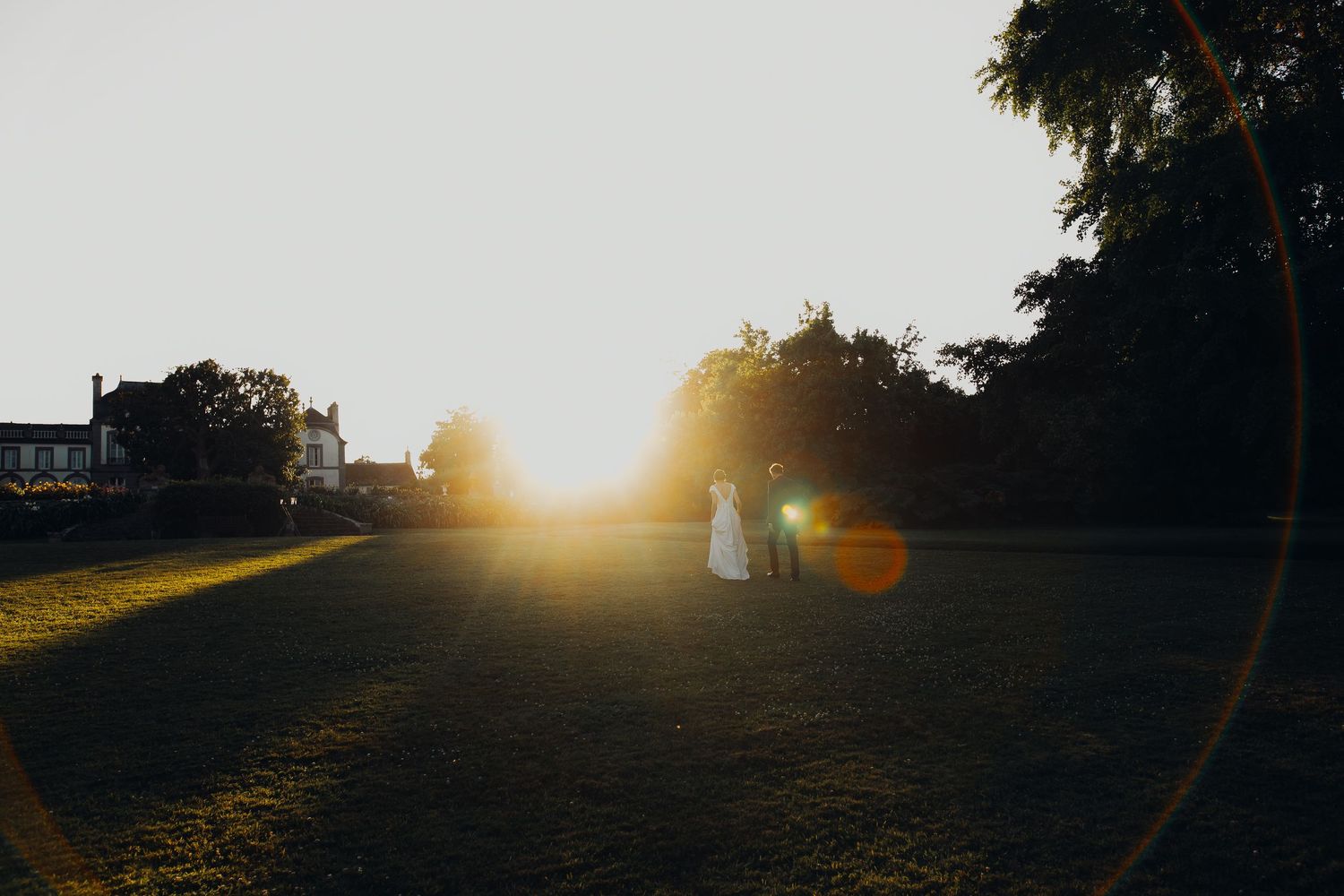 Bride and groom silhouetted against golden sunset light in open green field near historic estate.