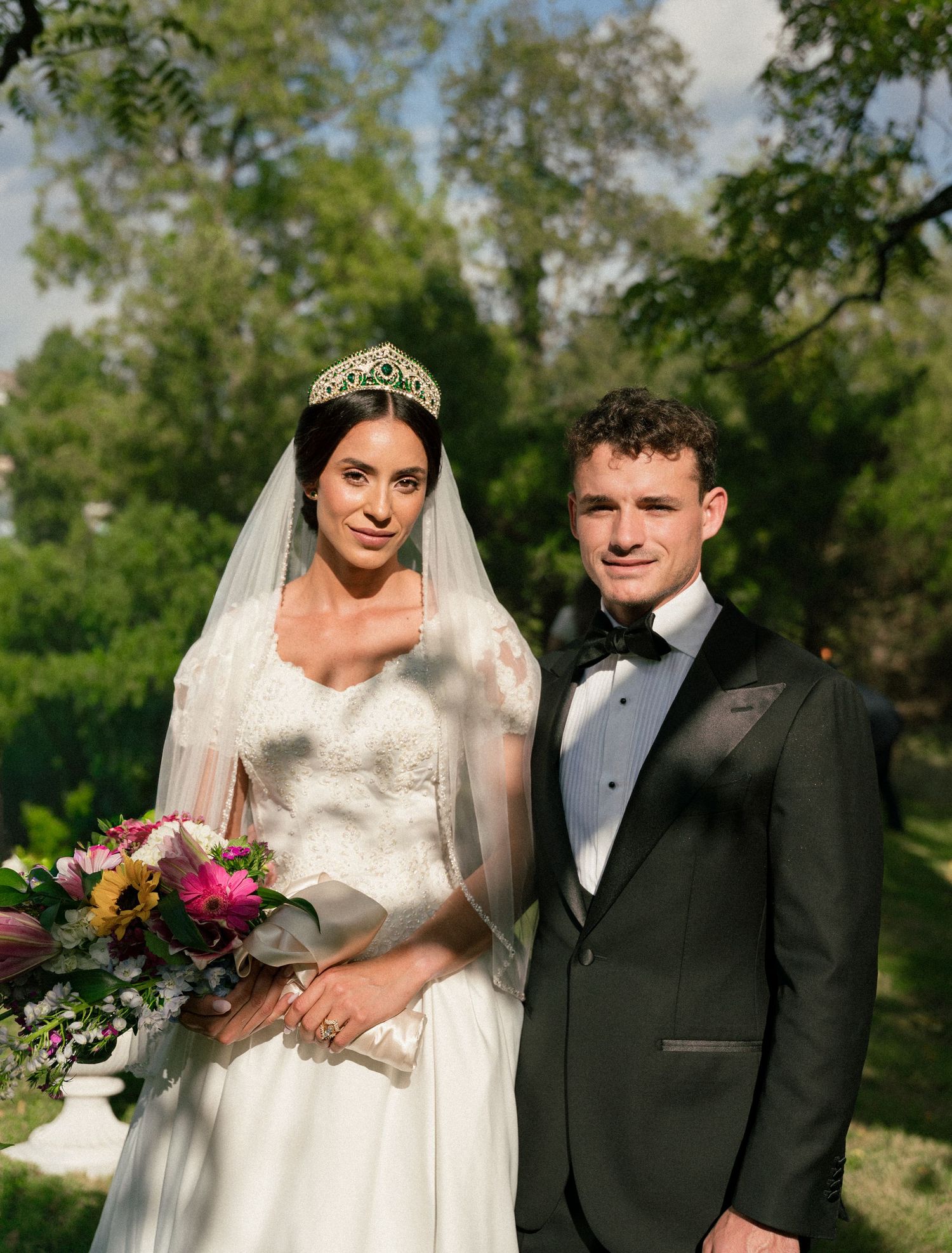 An outdoor wedding photoshoot featuring formal attire and vibrant floral bouquet against green trees.