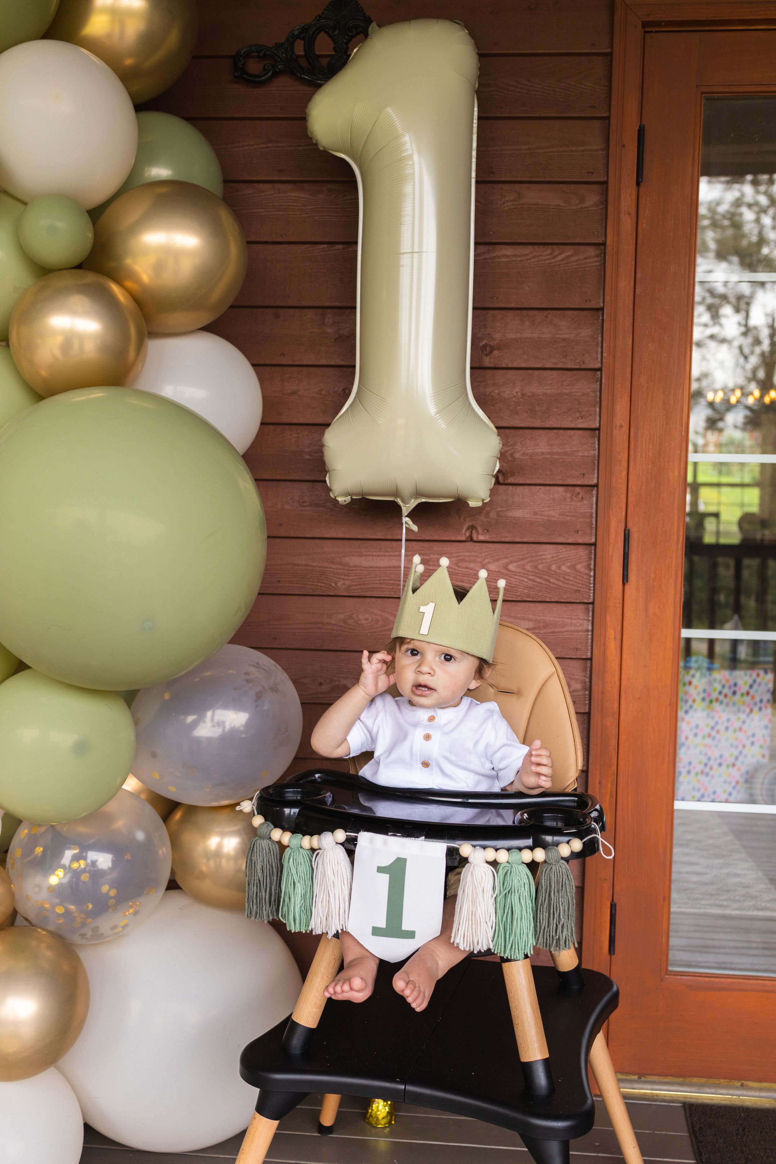 A baby in a white dress sits in a high chair decorated with sage green and gold balloons for their first birthday celebration.
