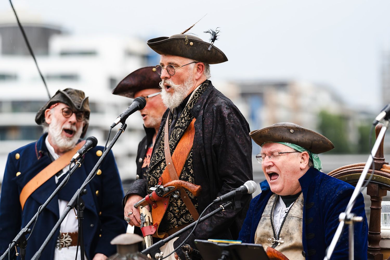 Three men in pirate costumes singing outdoors at a maritime festival, wearing tricorn hats and period clothing.