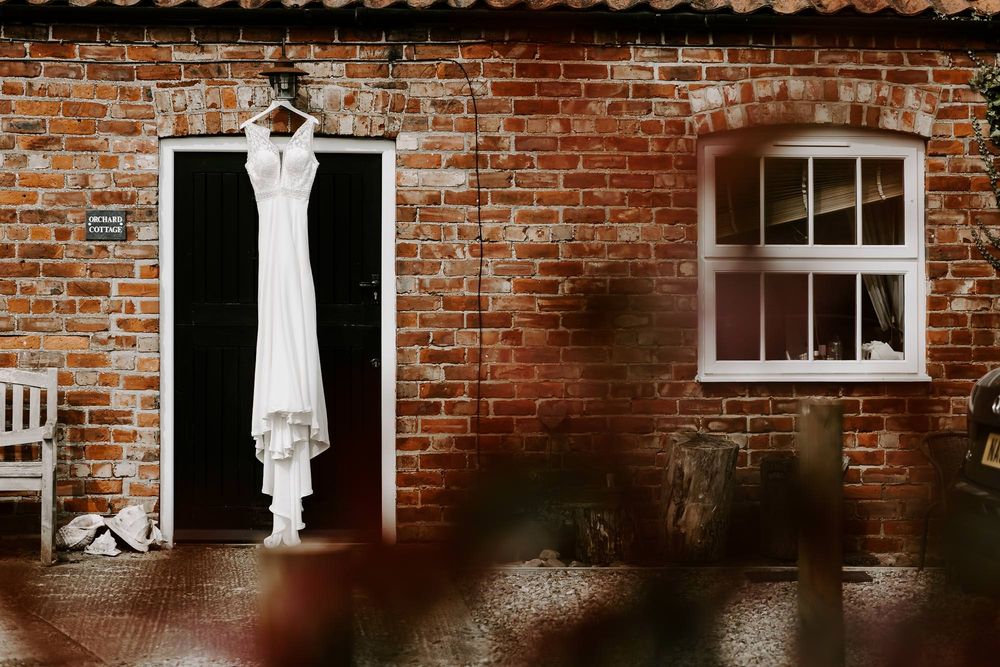 A white wedding dress hangs against a rustic brick wall with windows, creating an elegant contrast in natural light.