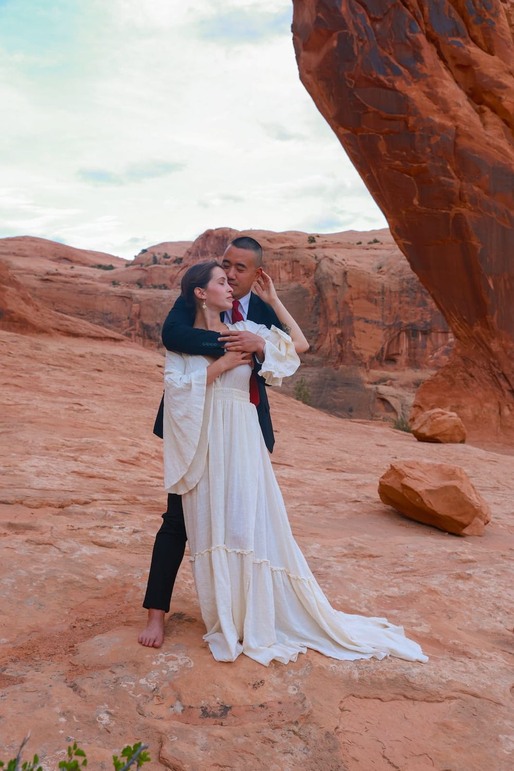 A couple shares an intimate moment under a red rock arch formation in a desert landscape.