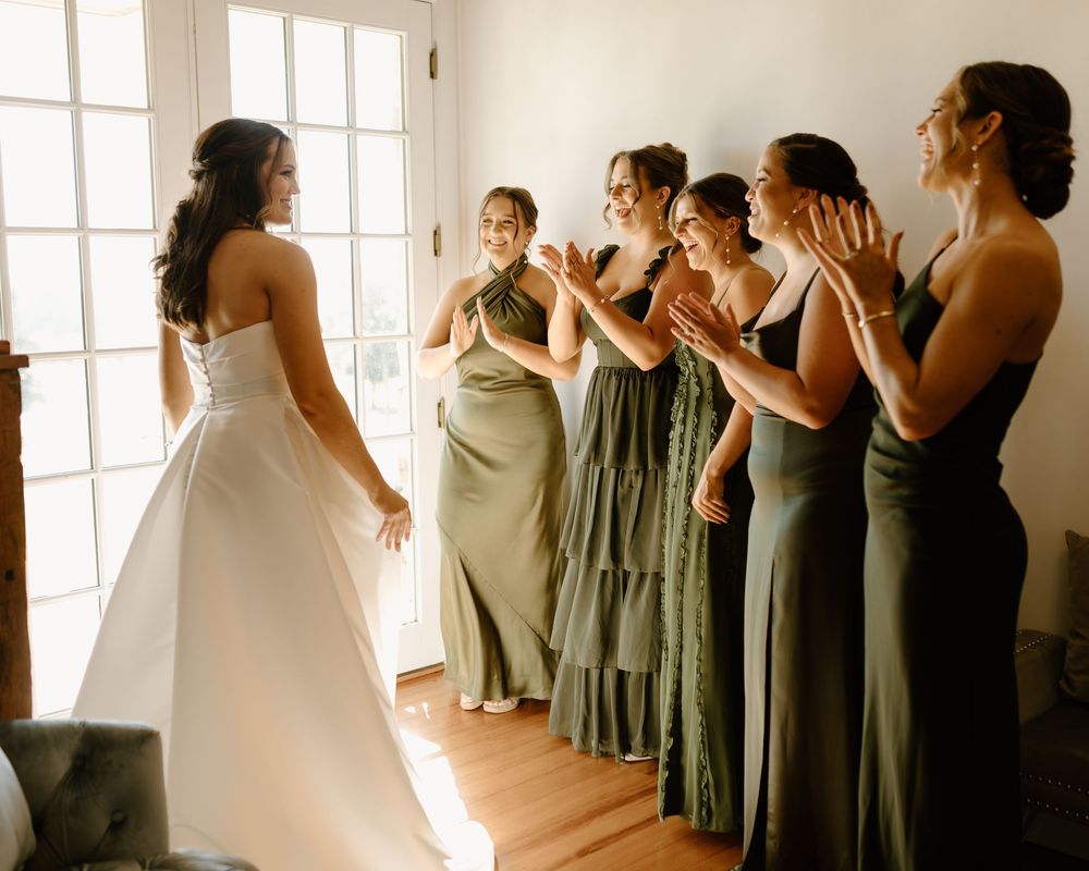 A group of bridesmaids in sage green dresses react emotionally as they see the bride in her wedding gown.