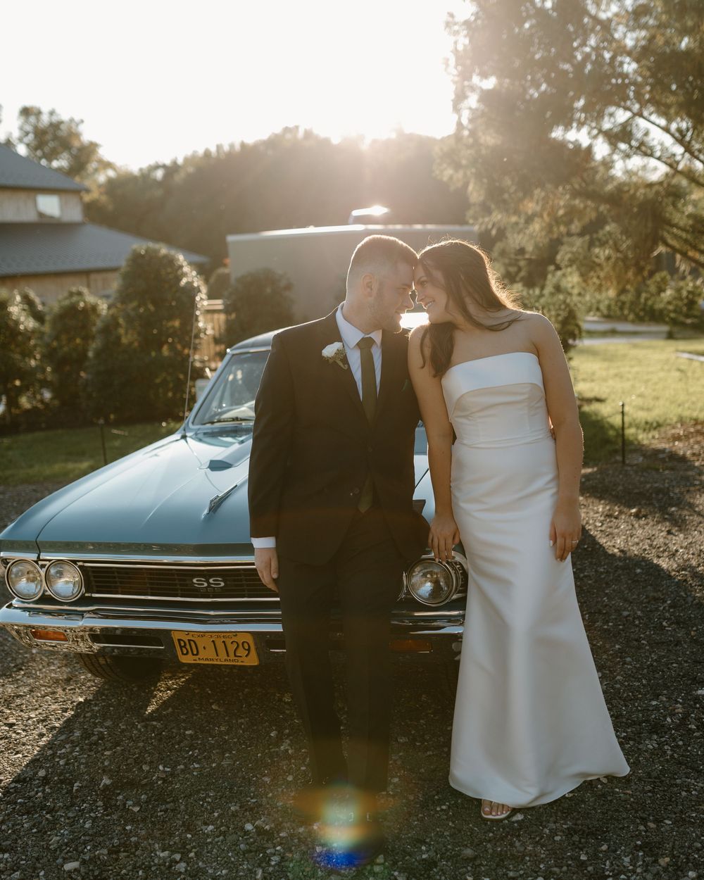 A couple poses beside a classic vintage car during sunset in a romantic outdoor setting.