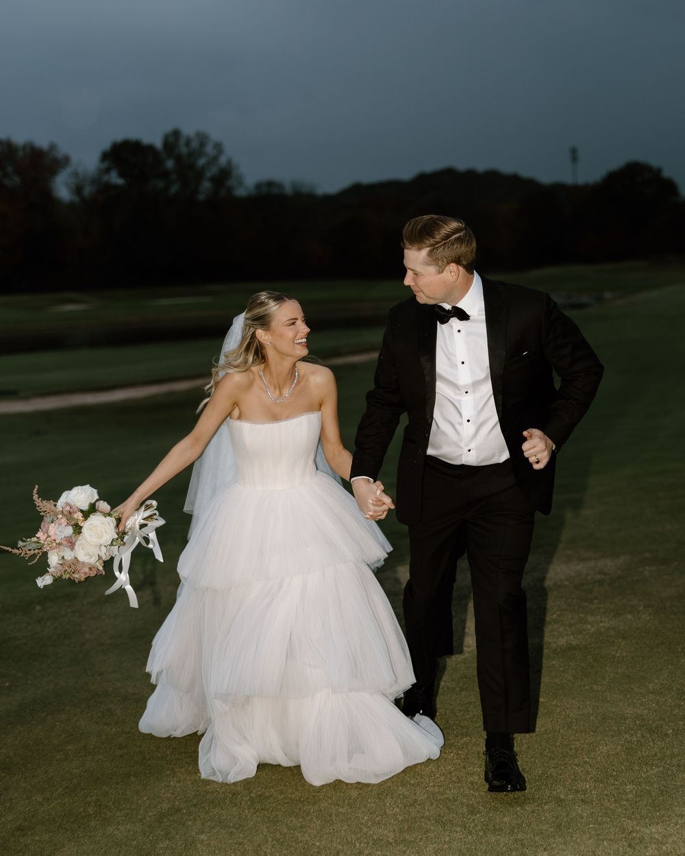 A couple in formal wedding attire playfully walks with their dog across a grassy field at dusk.