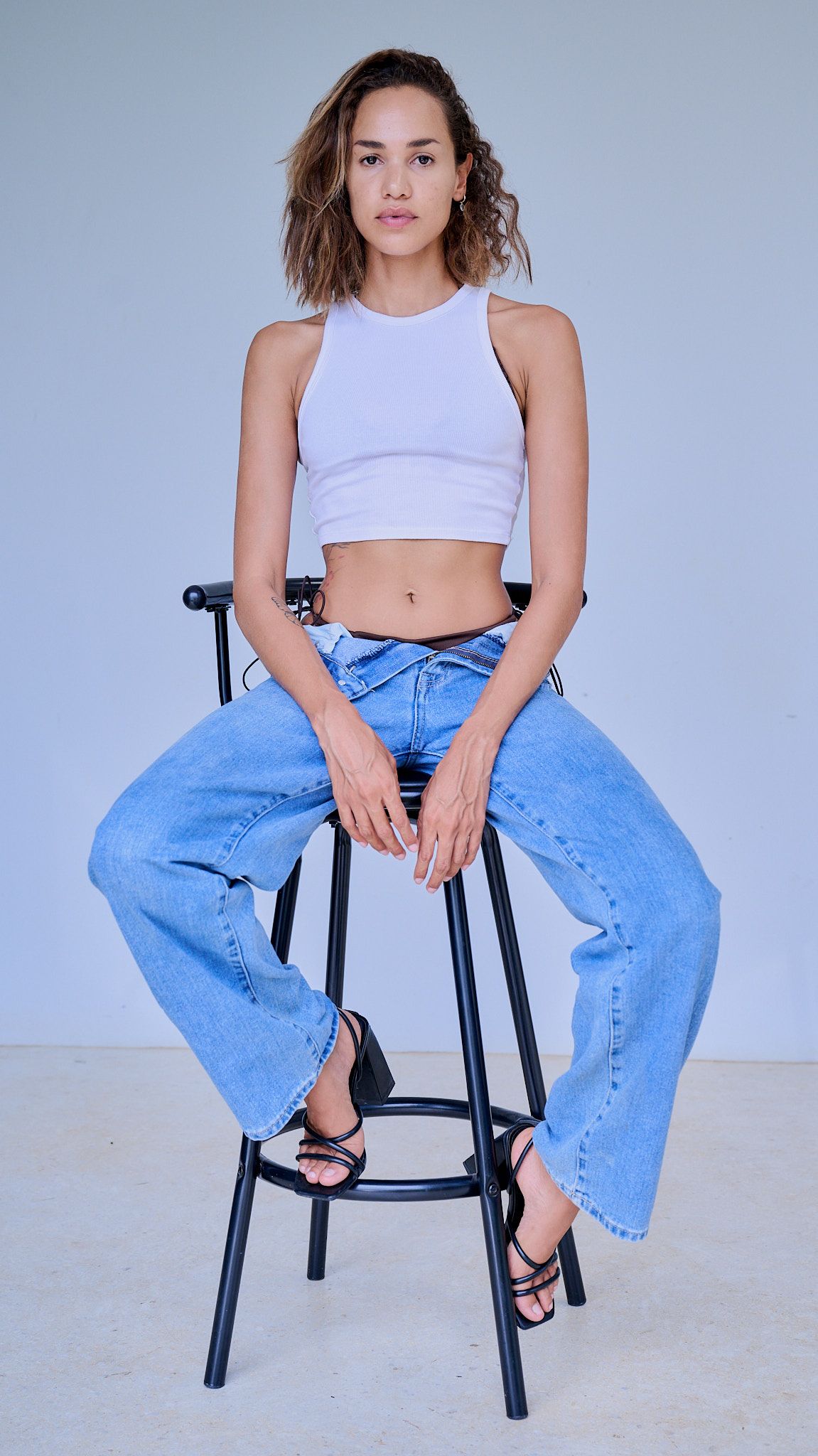 Model in white crop top and baggy blue jeans poses on black stool against light background in fashion photoshoot.