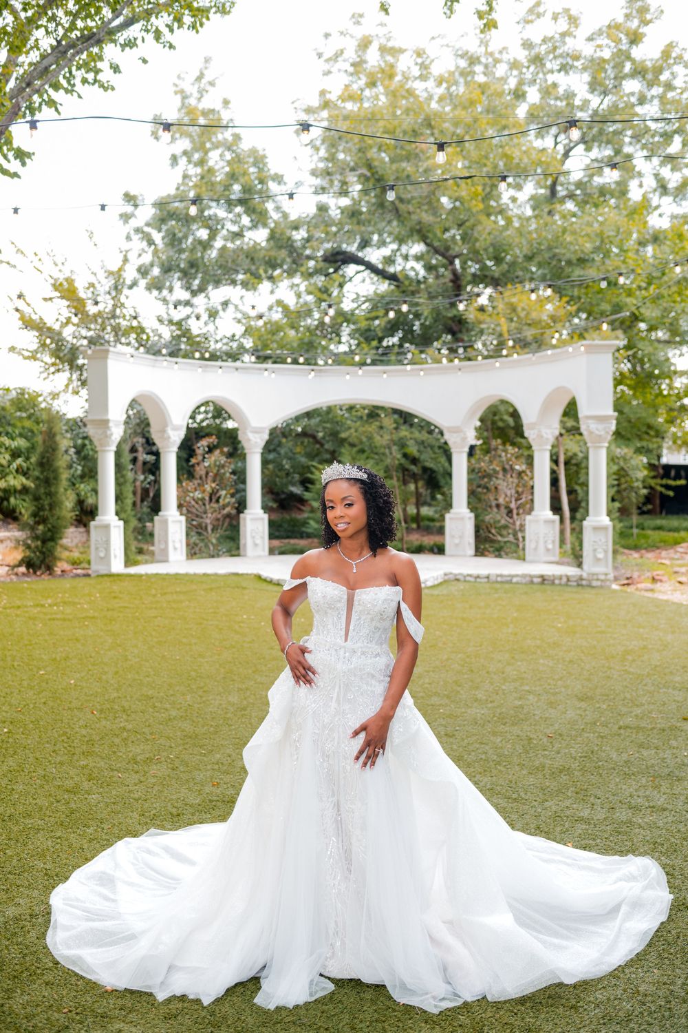 A flowing white wedding dress is elegantly displayed outdoors on a grassy lawn with white decorative arches in the background.