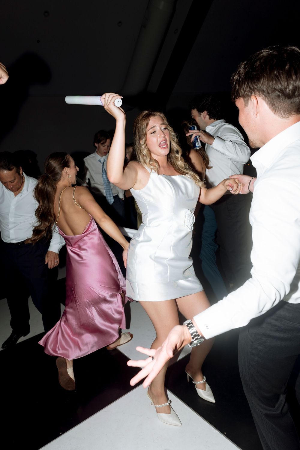 Partygoers dancing energetically on a dark dance floor with one person in a white dress and another in pink.
