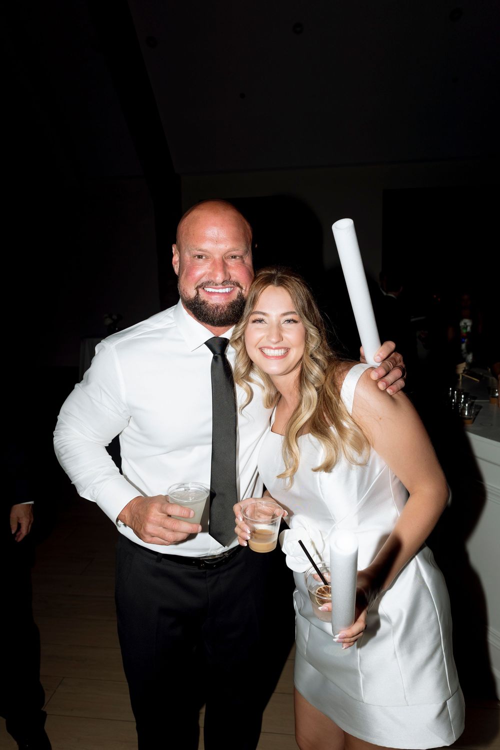 A couple in formal attire smiles while holding drinks at what appears to be a wedding reception or celebration.