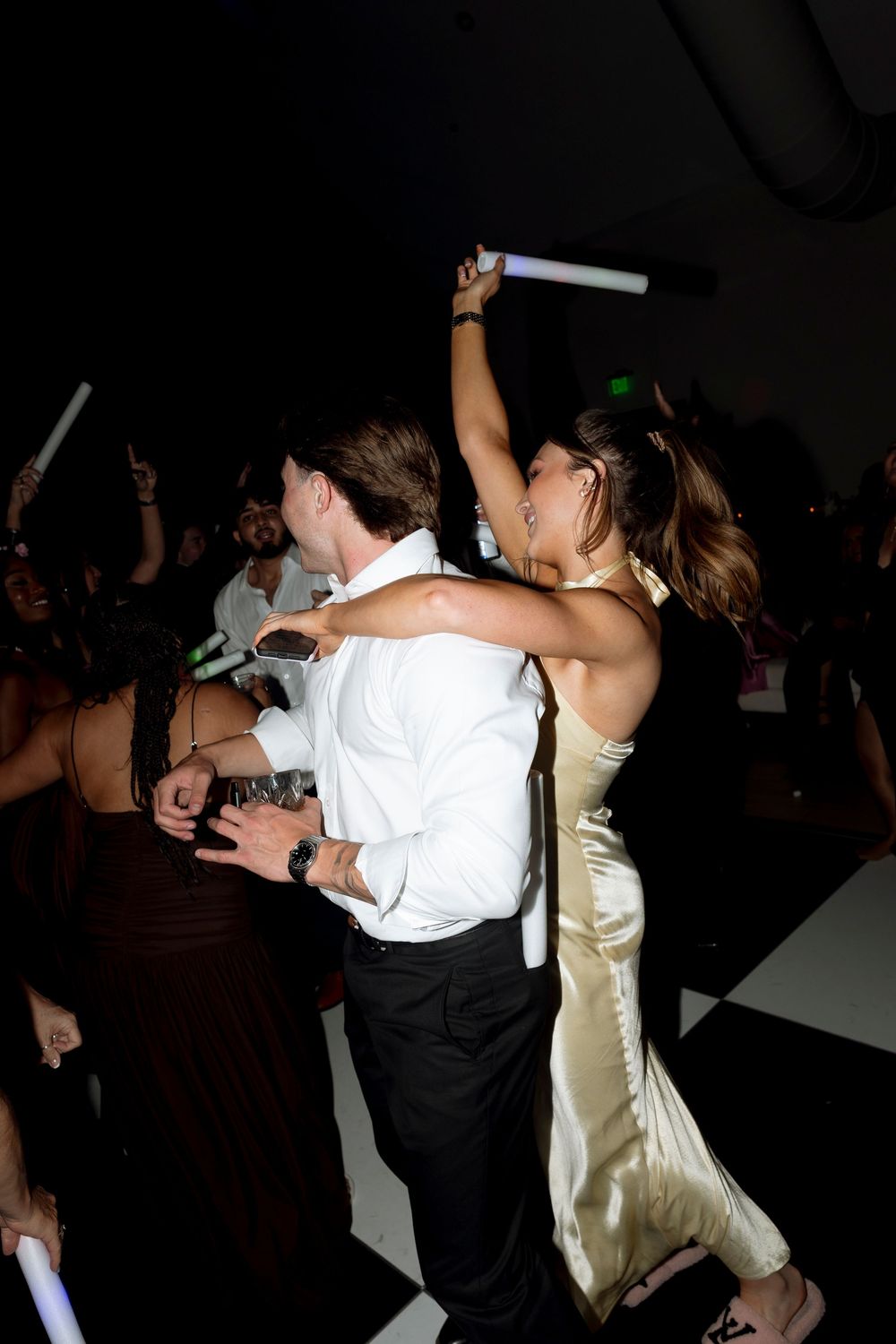People dancing together at a dark party venue with someone wearing a white shirt and gold dress visible.