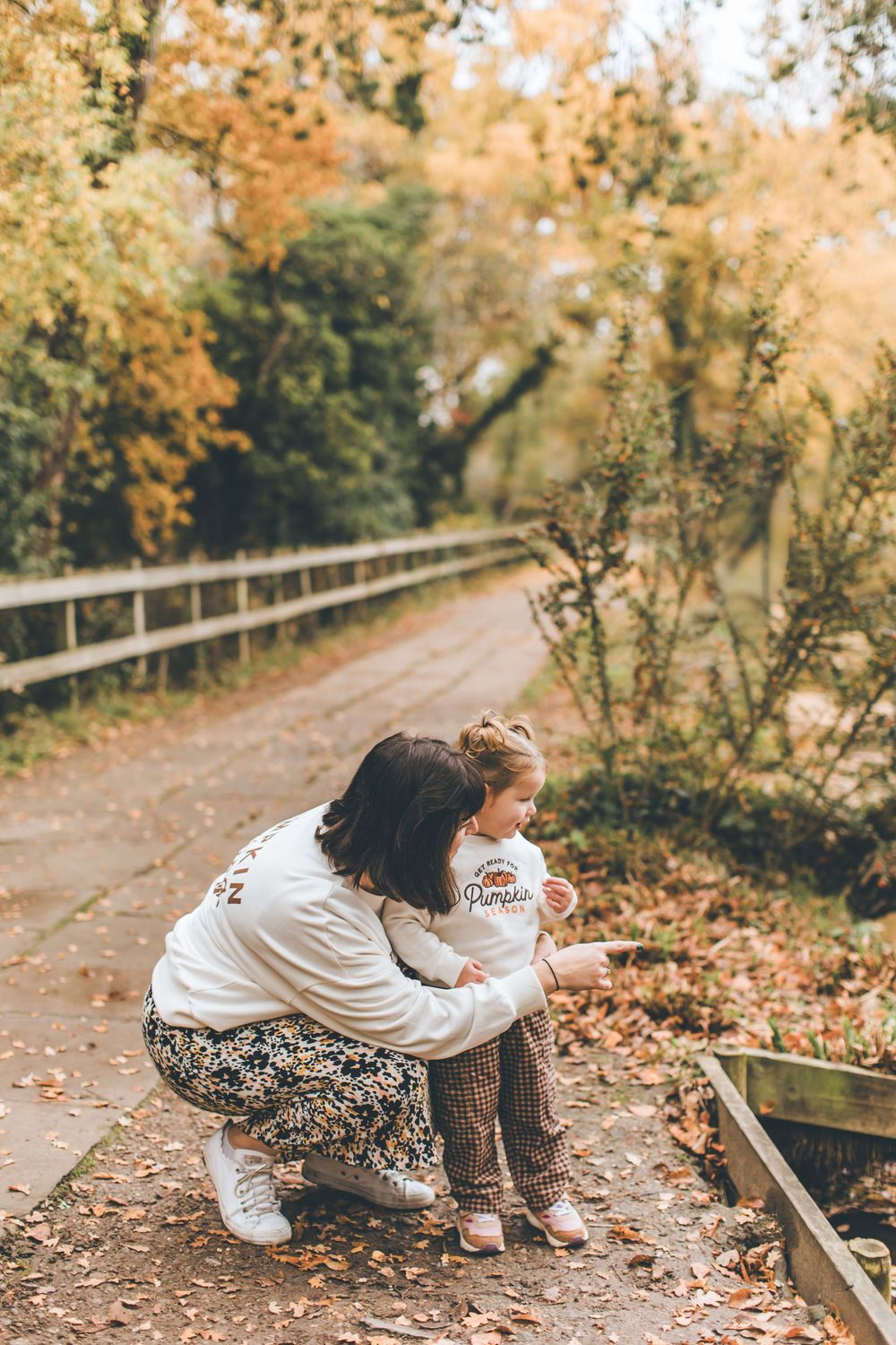 A person in a white sweater and patterned pants crouches down to explore fallen autumn leaves along a wooded path.