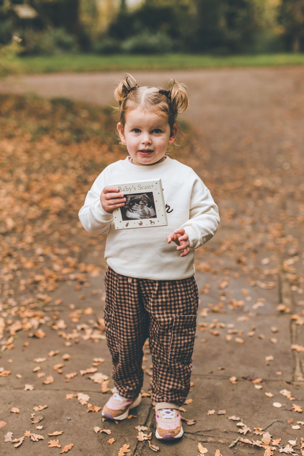 A child in a white sweater and plaid pants walks through fallen autumn leaves while holding a photograph.