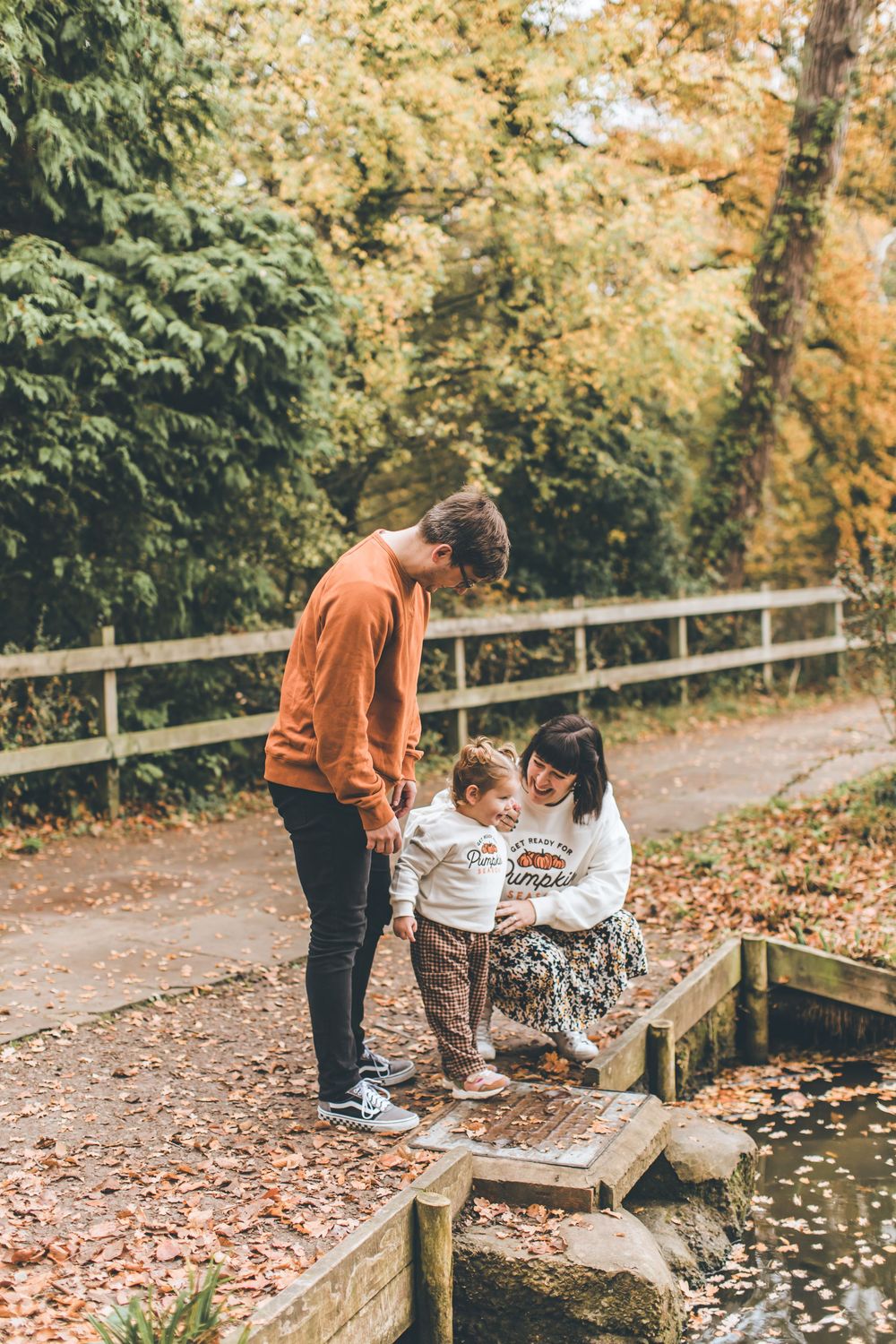 Two people explore a small pond or stream surrounded by fallen autumn leaves and wooden railings.