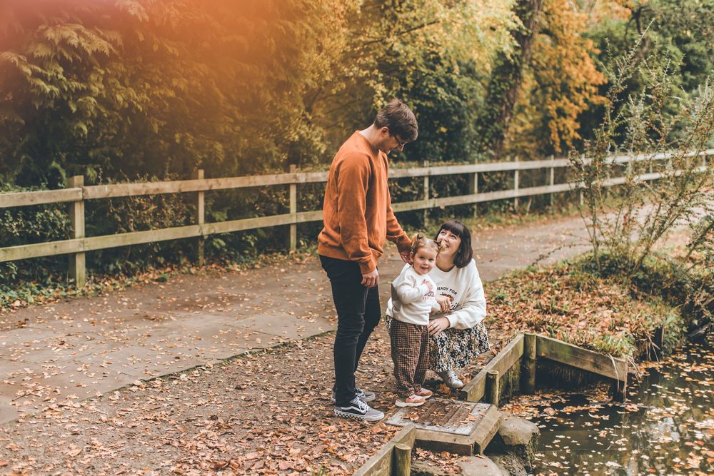 A family walks together on a wooden bridge surrounded by autumn foliage in a park setting.