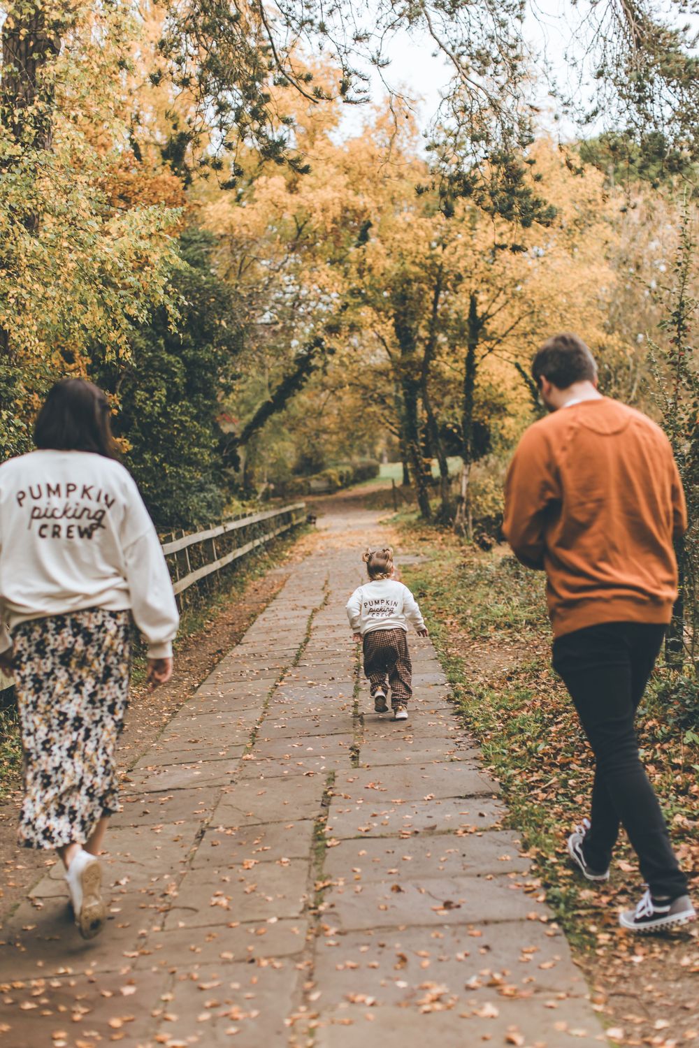 People walk along a leaf-covered path through an autumn forest wearing seasonal clothing.