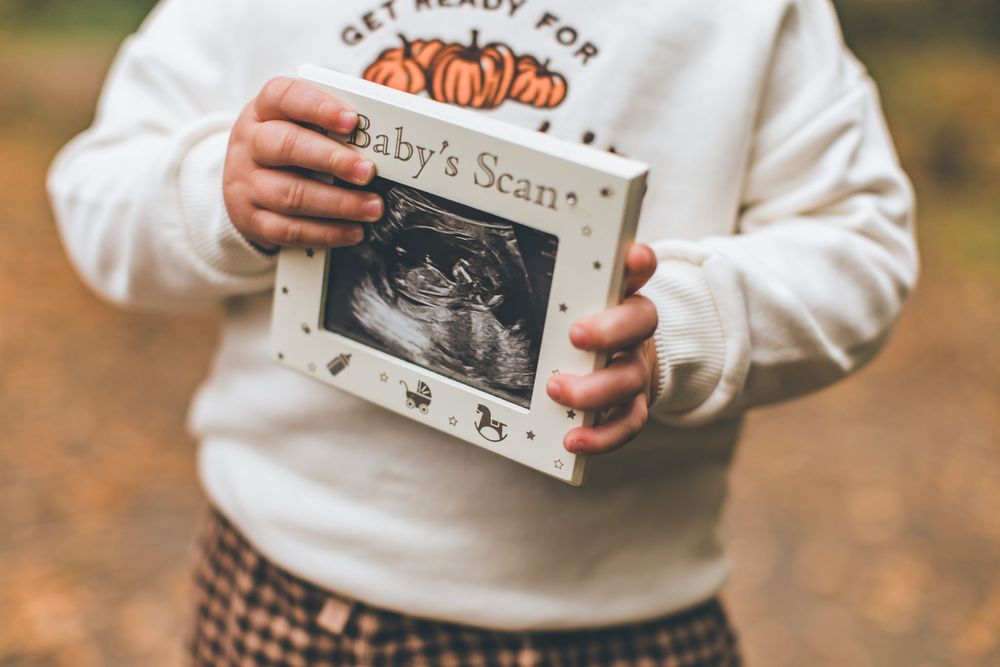 Hands holding a baby scan ultrasound photo frame against autumn background with plaid skirt and white sweater.