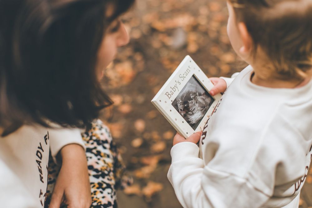 Person in white sweater holds black and white ultrasound photo while sitting on autumn leaves.