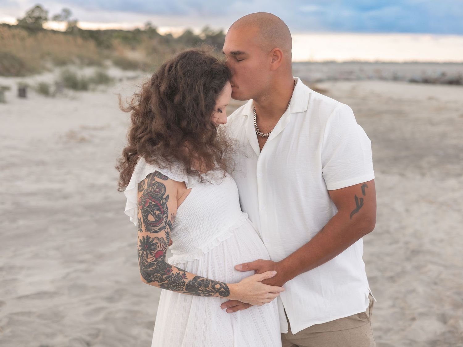 A romantic couple embraces on a sandy beach, with the person on the left wearing a white dress and displaying arm tattoos.