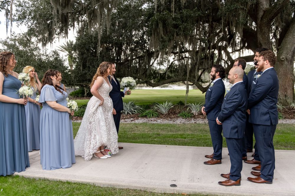 Wedding party in light blue dresses and dark suits poses together on stone pathway in outdoor garden setting.