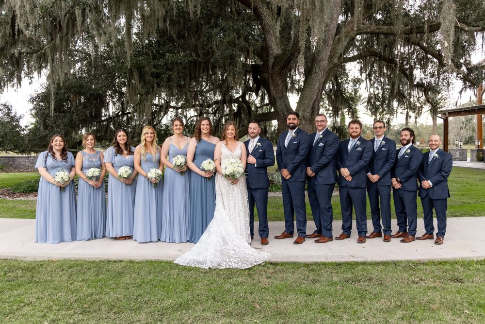 Wedding party in blue and navy attire poses together on garden path with Spanish moss trees in background.