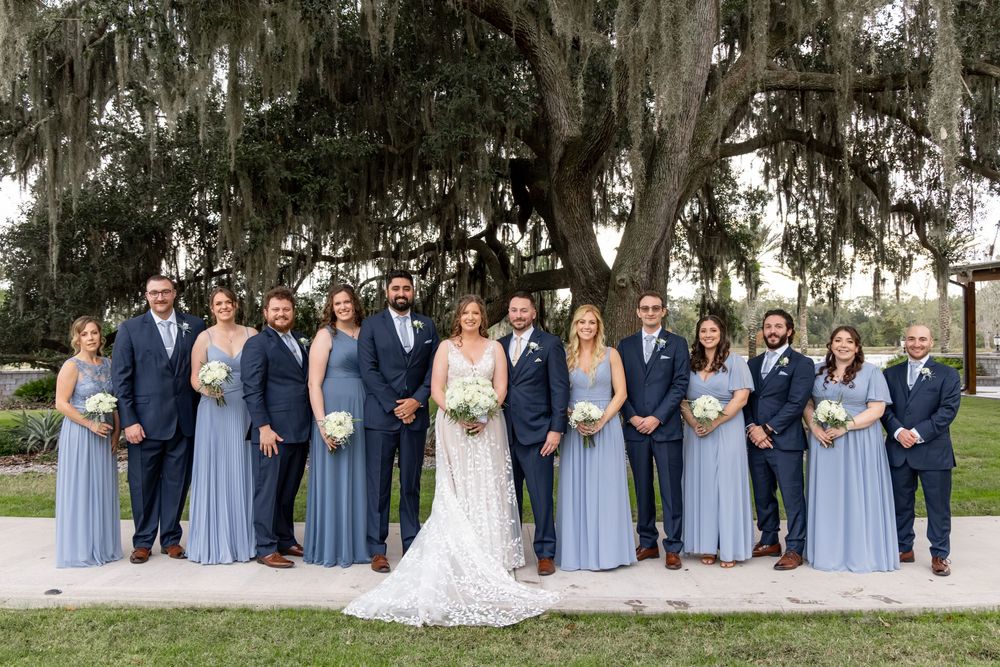 Wedding party in blue and navy attire poses together under moss-draped oak trees in an outdoor setting.