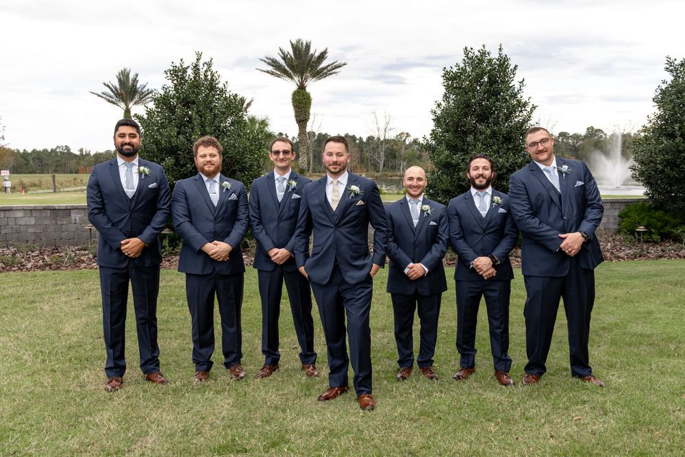 Groomsmen in navy suits and brown shoes sit casually on grass for wedding party photos.