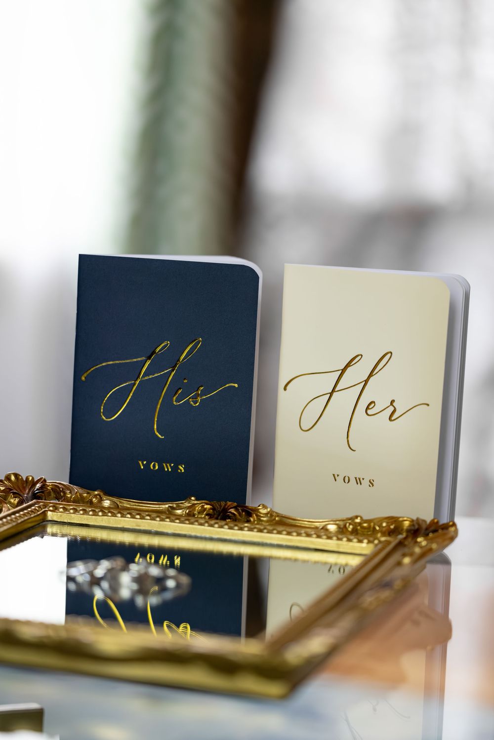 Navy blue and gold wedding vow books with 'His' and 'Her' text displayed on an ornate gold tray.