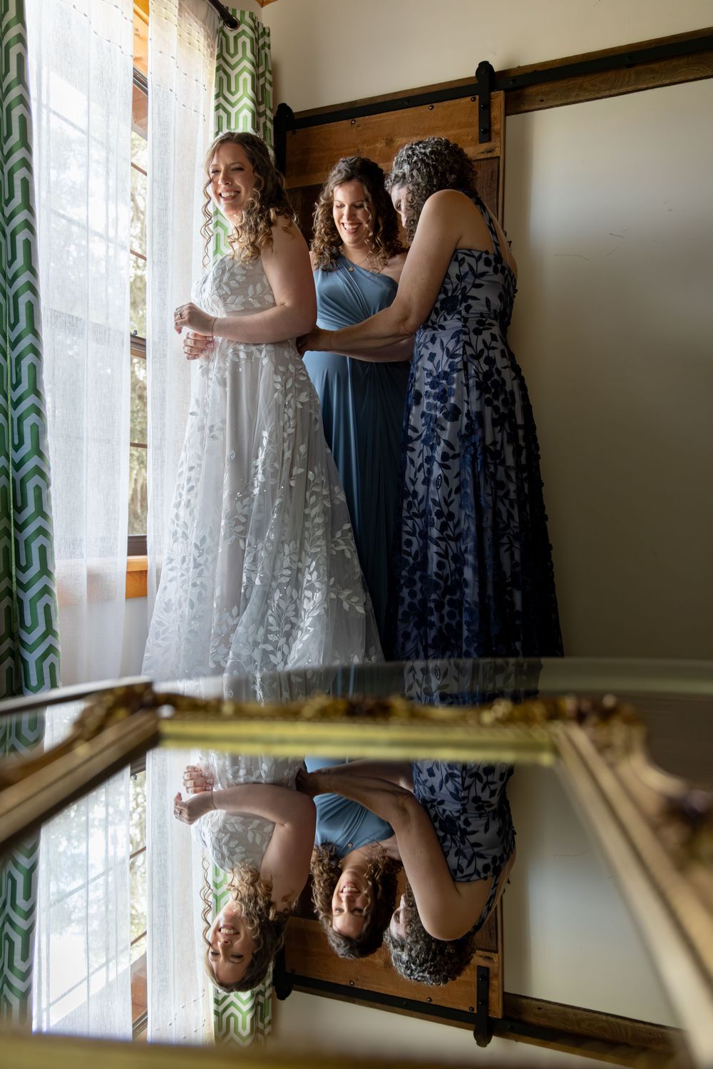 Three women in formal dresses share a candid moment reflected in a mirror during wedding preparations.