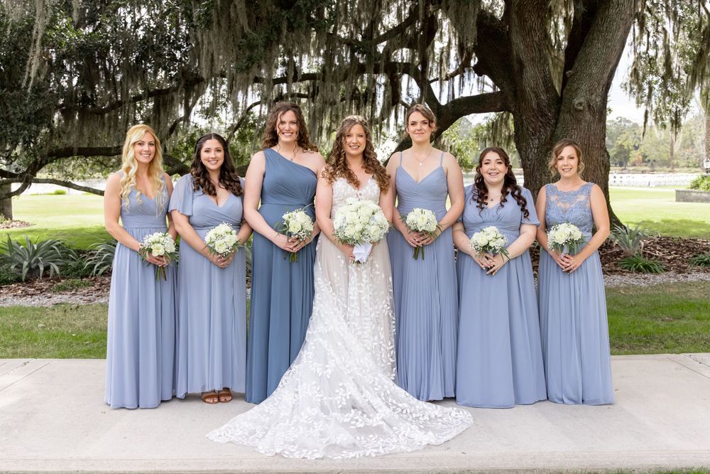 A wedding party in light blue bridesmaid dresses and white bridal gown poses together in an outdoor garden setting.