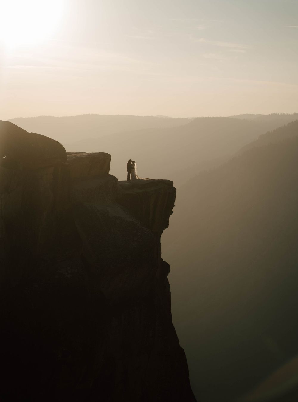 Silhouette of couple embracing on dramatic cliff edge at golden sunset in Yosemite.