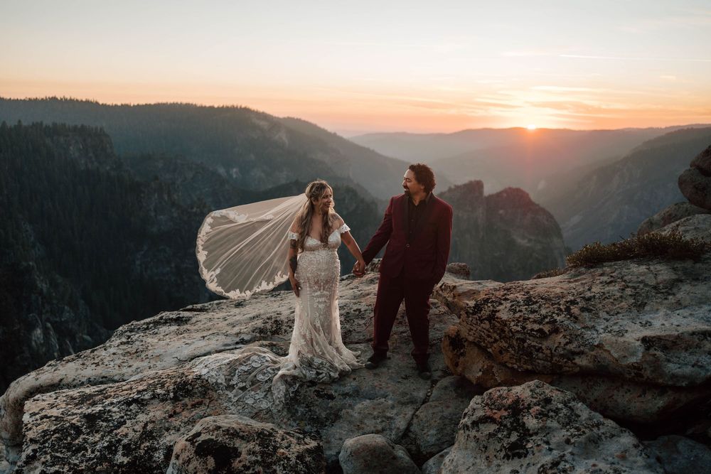 Bride and groom holding hands on cliff edge at sunset with stunning Yosemite valley panoramic view.