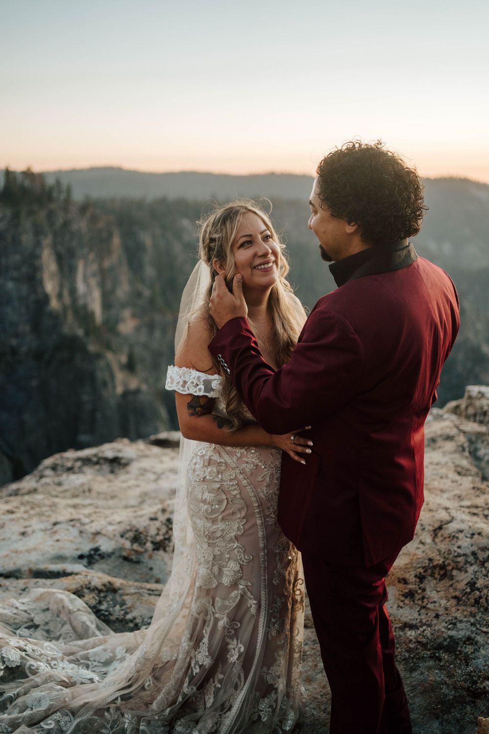 Groom in burgundy suit tenderly touching brides face as she smiles during mountain sunset elopement.