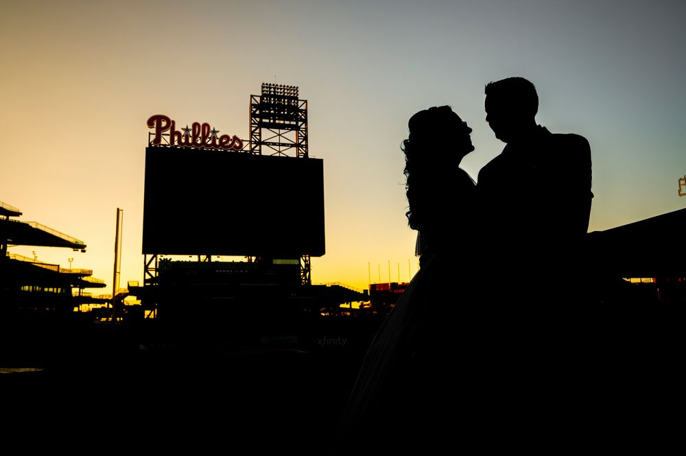 Silhouettes of baseball stadium structures and spectators against a dramatic sunset sky during a game.