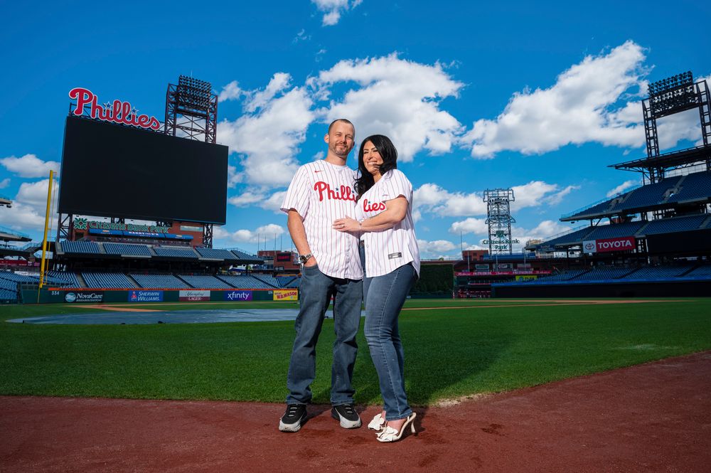 A couple wearing Philadelphia Phillies jerseys poses together on the baseball field at Citizens Bank Park on a sunny day.
