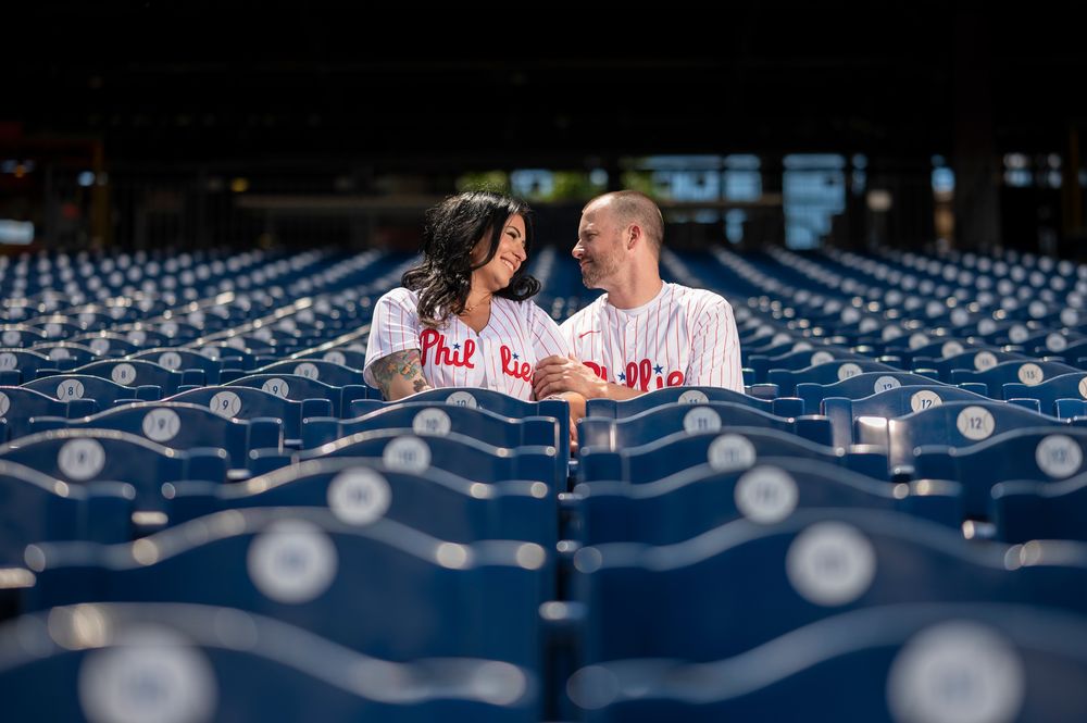 A couple shares a romantic moment in an empty baseball stadium with blue seats and string lights overhead.