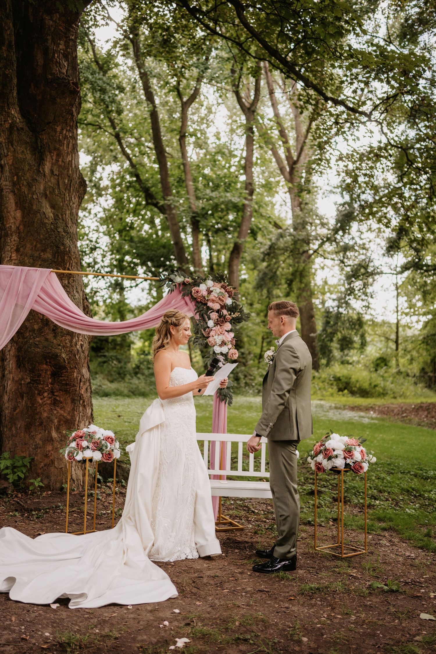 A romantic outdoor wedding ceremony in a wooded setting with pink and floral decorations and elegant white seating.