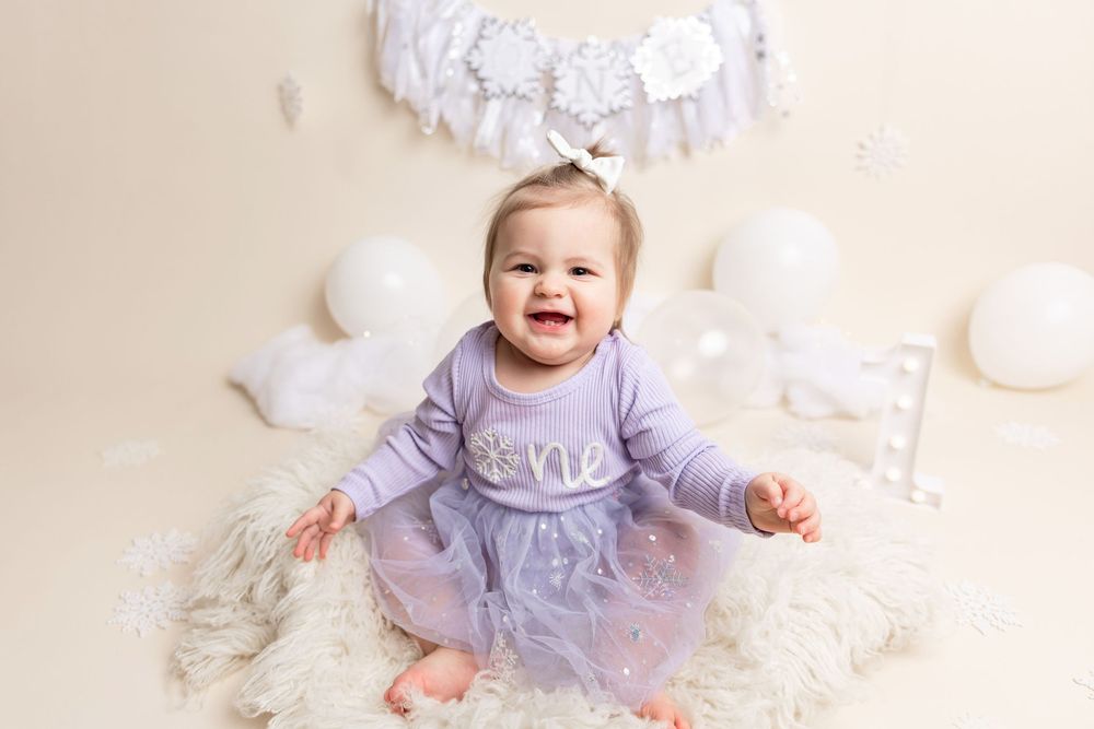A small child wearing a lavender dress sits on a white fluffy rug with a joyful expression.