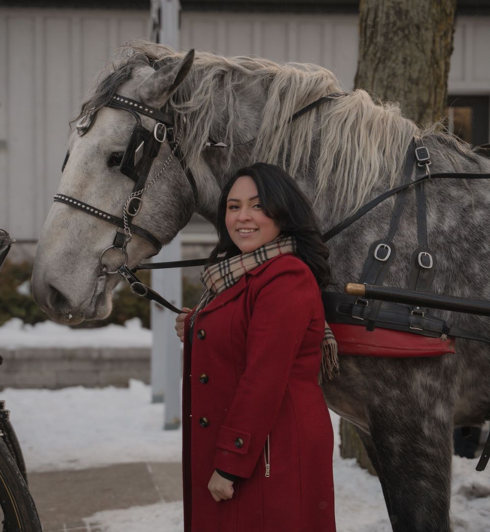 A person in a red winter coat stands next to two horses in a snowy setting.