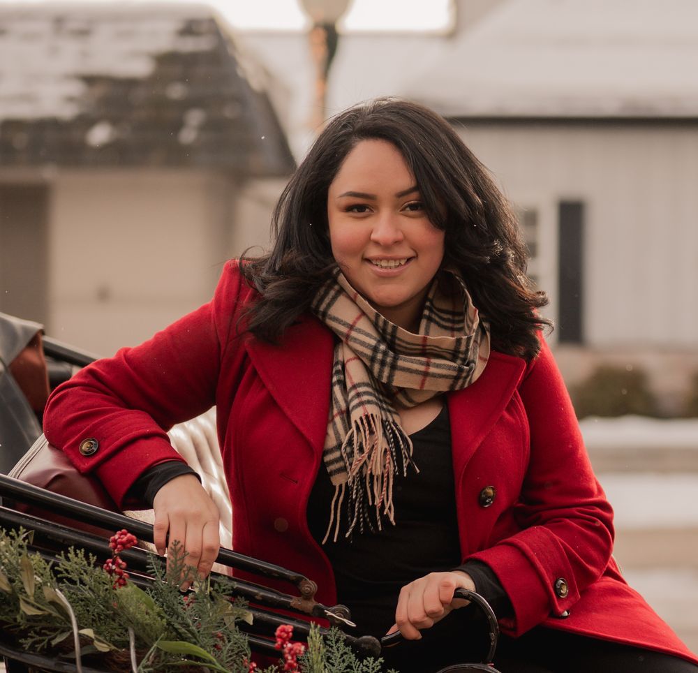 A person in a red coat and plaid scarf works on decorating an evergreen wreath outdoors during winter.