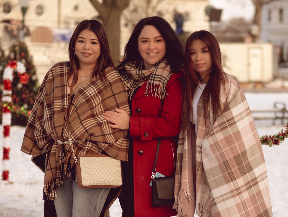 A group in winter outfits poses together wearing plaid shawls and red coats during a snowy outdoor photoshoot.