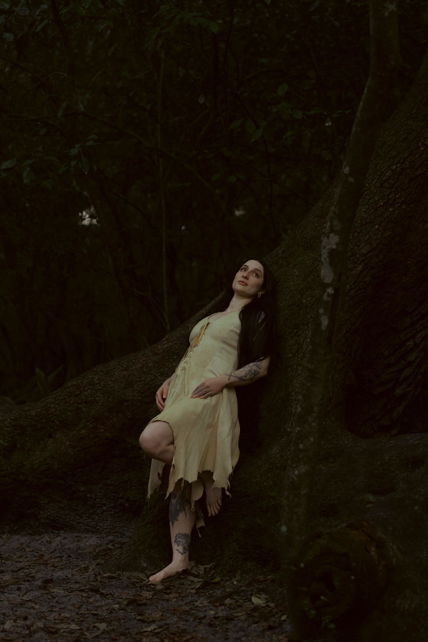 A person in a light dress leans against a mossy tree trunk in a dark, moody forest setting.