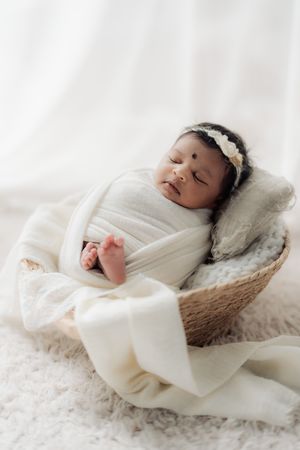 Newborn sleeping in cozy cream blanket inside woven basket on white textured backdrop.