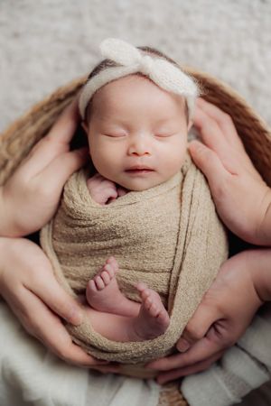 Sleeping newborn wrapped in beige fabric nestled in parents' hands.