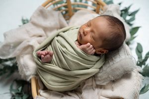 Sleeping newborn in sage green wrap on cream blanket with greenery accents.