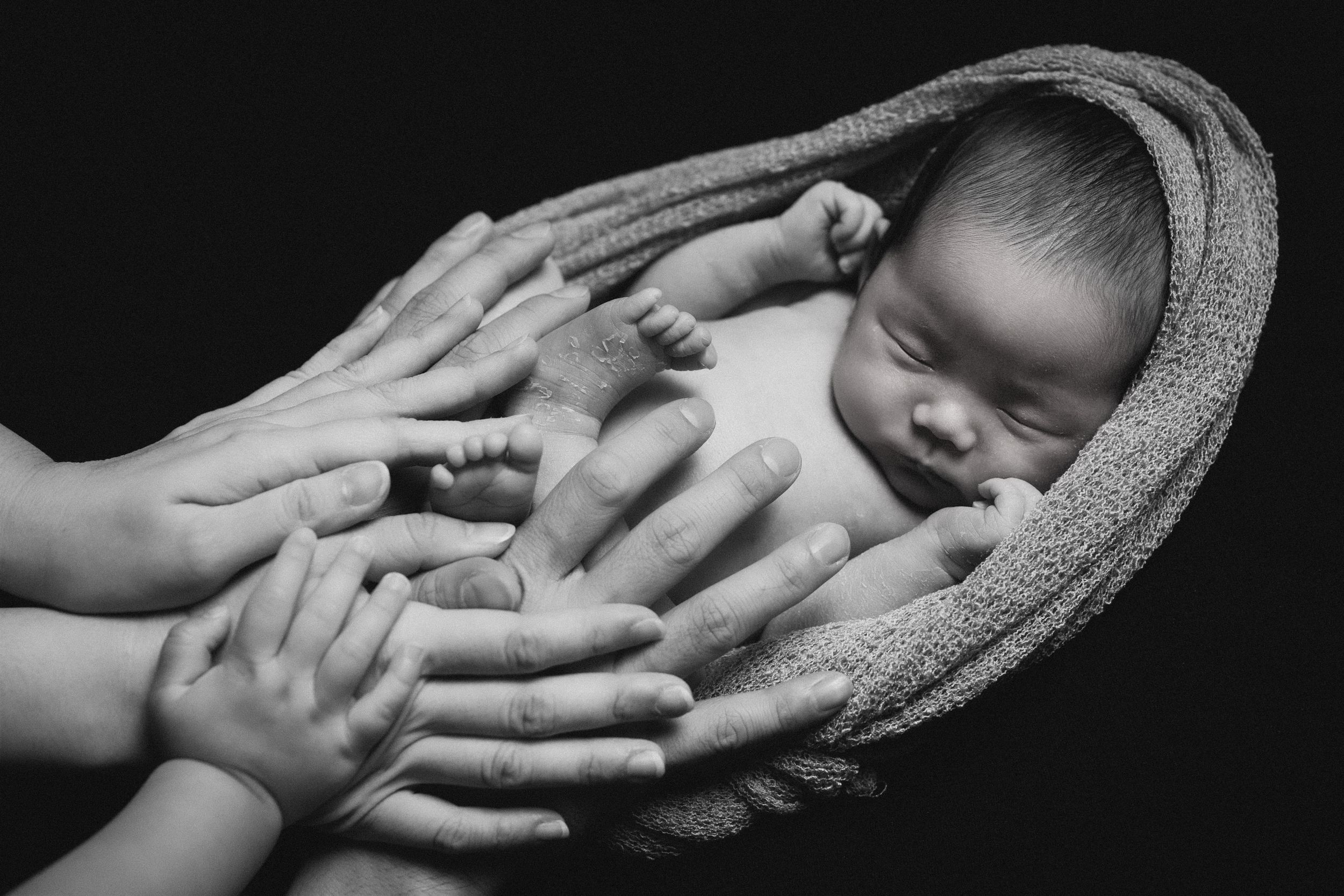 Gentle hands cradle sleeping newborn baby wrapped in textured blanket in monochrome.