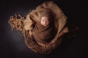 Newborn photography featuring a tiny baby nestled in a brown woven basket against dark background.