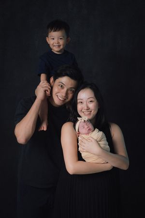 A family portrait against a dark background showing parents with their toddler and newborn baby.
