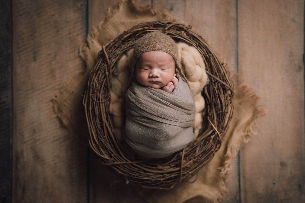 Newborn baby wrapped in soft fabric nestled in rustic wooden nest against textured background.